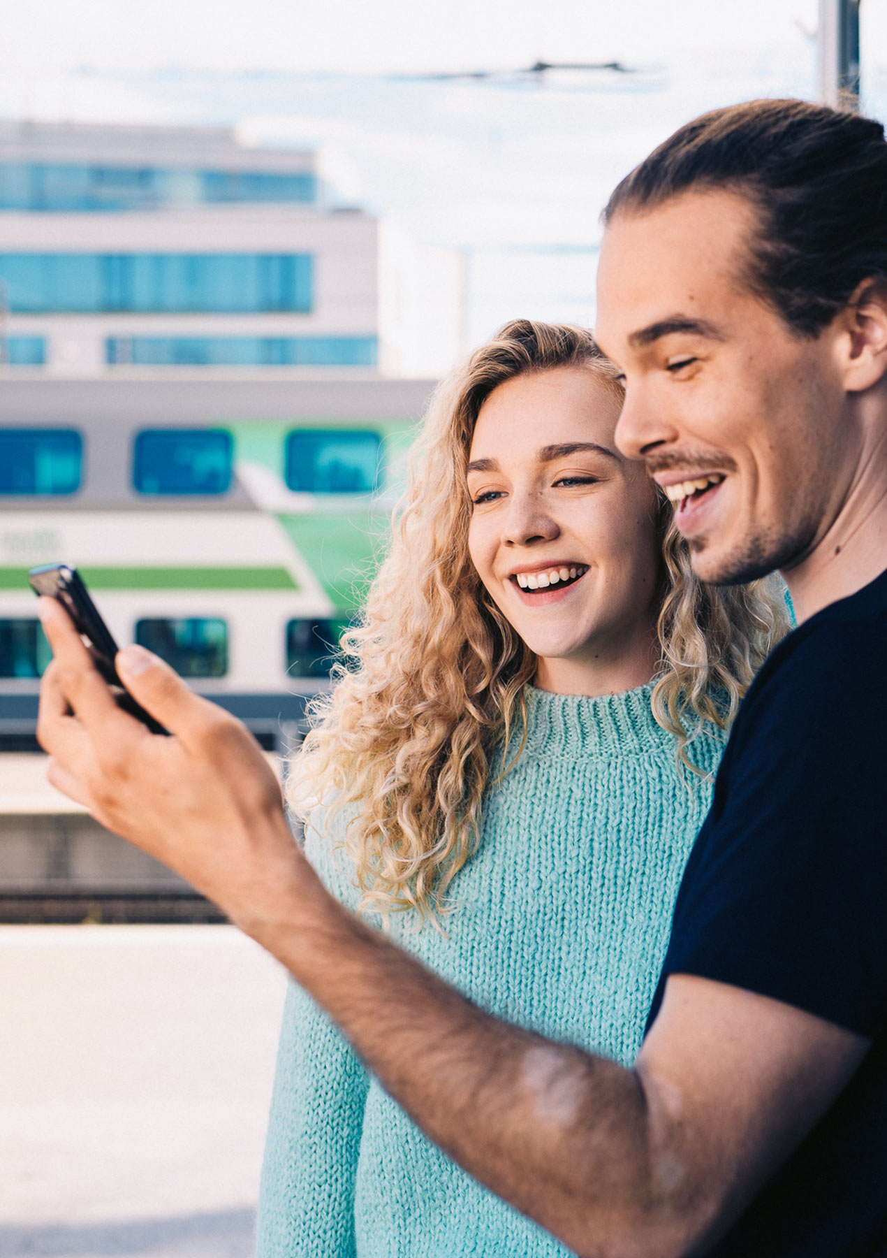 Woman and a man in the railway station. The man is holding a mobile phone and both of them are looking at it.