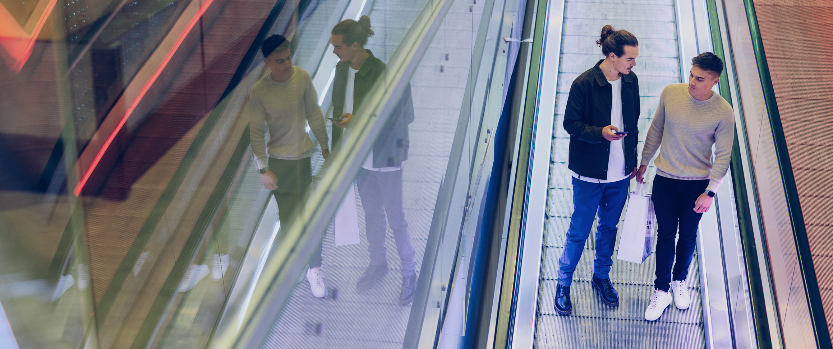 Two men standing in escaltor chatting. One of them is holding a phone.