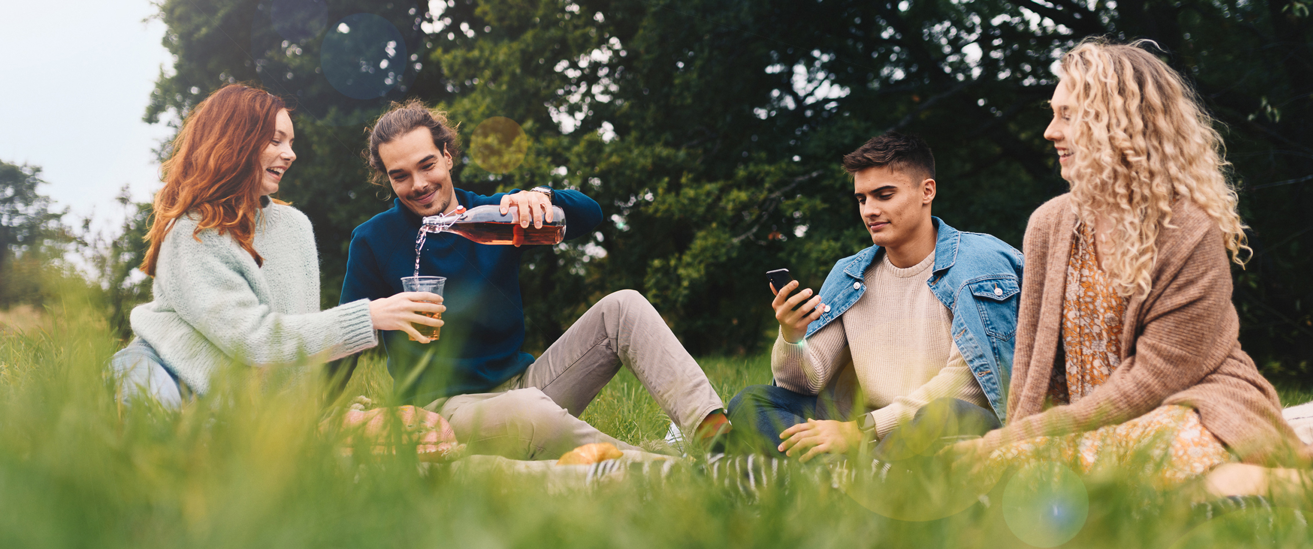 People outside in the park having a picnic. One man is holding a phone and looking at it.
